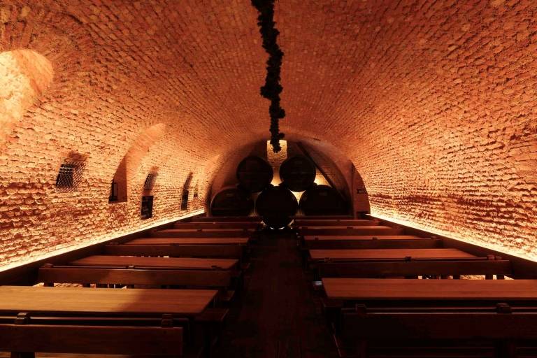 In a large vaulted brick cellar, several tables and beer benches are lined up, and a number of wooden barrels are stacked up