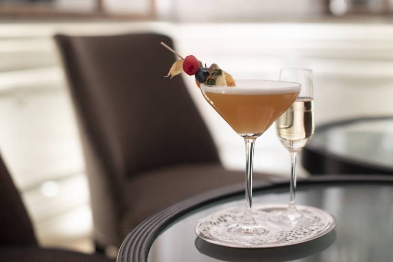 A cocktail and a champagne glass on a small silver tray, in the background a blurred chair in Falk's Bar in Munich.