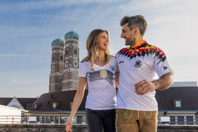 Zwei Fußballfans tragen Deutschlandtrikots in München und im Hintergrund sind die Türme der Frauenkirche zu sehen.