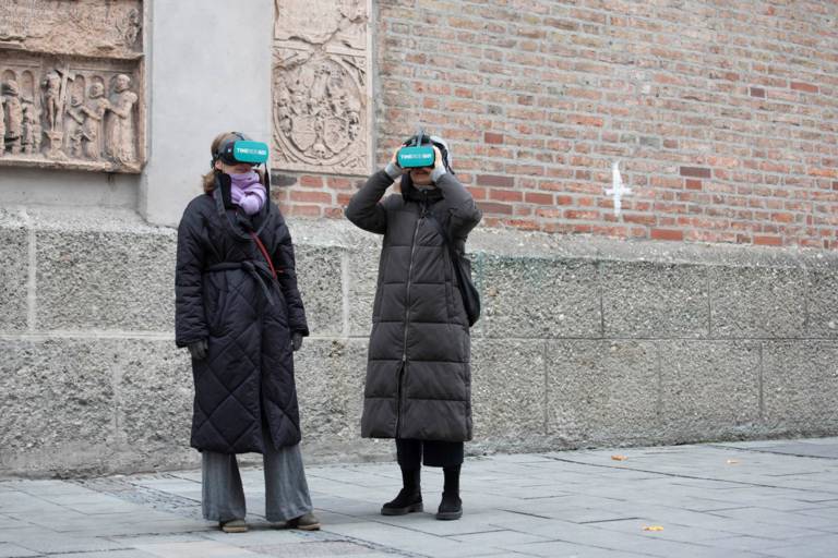 Zwei Frauen in Wintermäntel stehen  mit VR-Brille vor der Münchner Frauenkirche