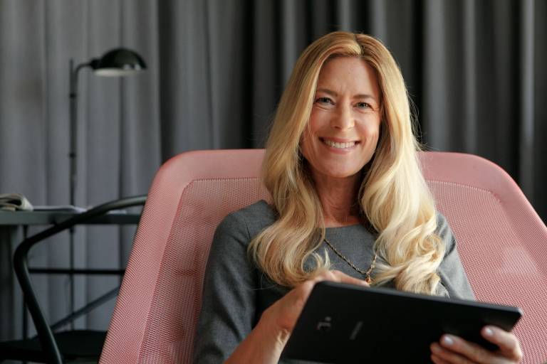 Eine Frau sitzt in einem Sessel und schaut lächelnd in die Kamera während sie ein Tablet in der Hand hält.
