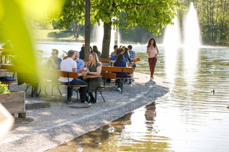 Mehrere Menschen sitzen in einem Biergarten in München am Wasser und unterhalten sich.