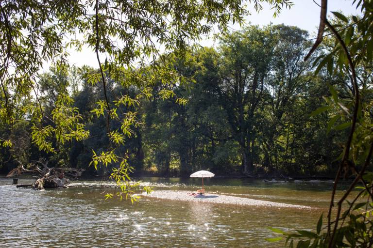 Eine Person liegt unter einem Sonnenschirm auf einer Sandbank in der Isar in München.