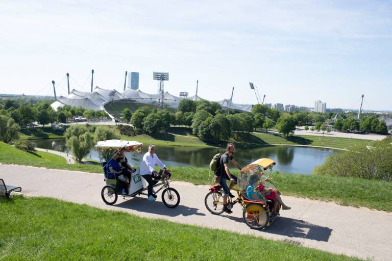 Ein Mann fährt mit seiner Rikscha und Gästen durch den Olympiapark in München.