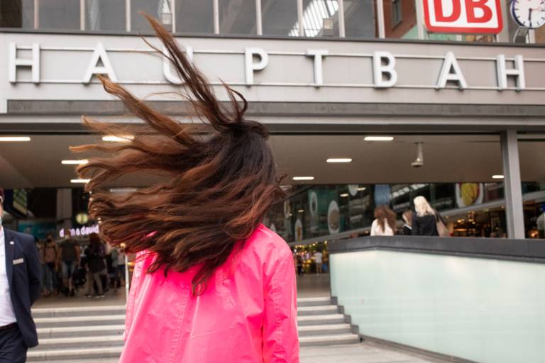 Eine Frau mit pinkem Regenmantel und fliegenden Haaren geht auf den Eingang eies Hnauptbahnhofs zu.