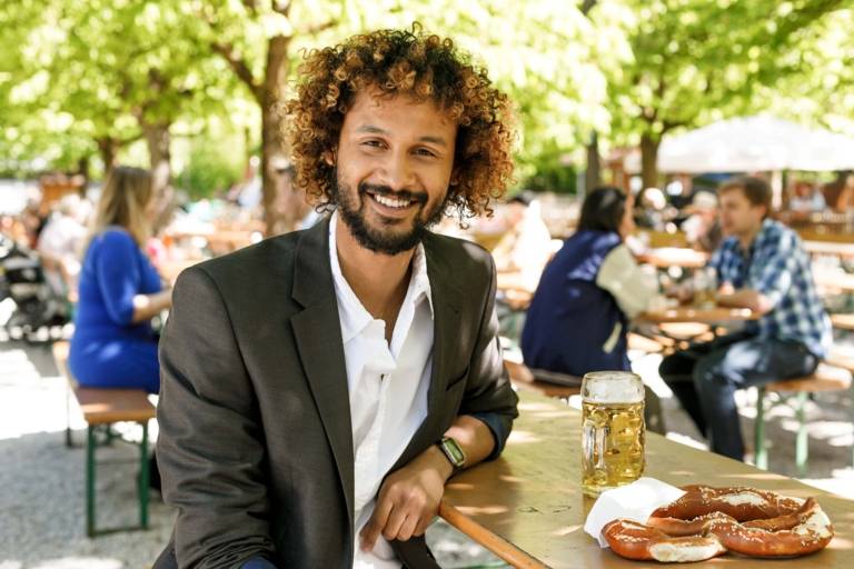 Un uomo con i capelli ricci e una giacca è seduto con pretzel e birra a un tavolo in una birreria all'aperto a Monaco.