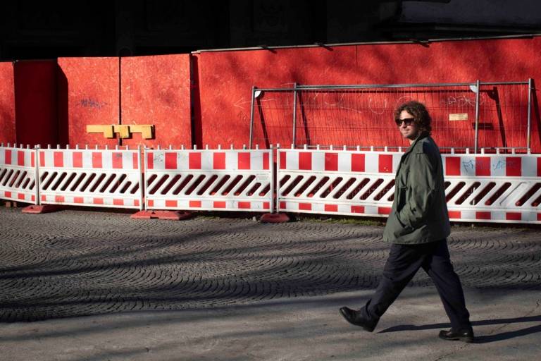 A man walks along a red wall and smiles at the camera.