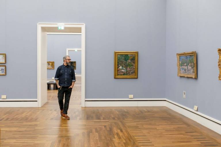 Ein Mann mit Brille und Bart geht durch die Neue Pinakothek in München und betrachtet die Gemälde.